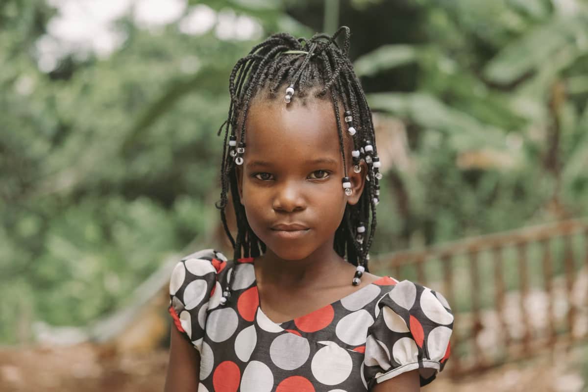 Haïti : Une fille se tient debout dans une robe à motifs. Elle a des tresses avec des perles blanches.