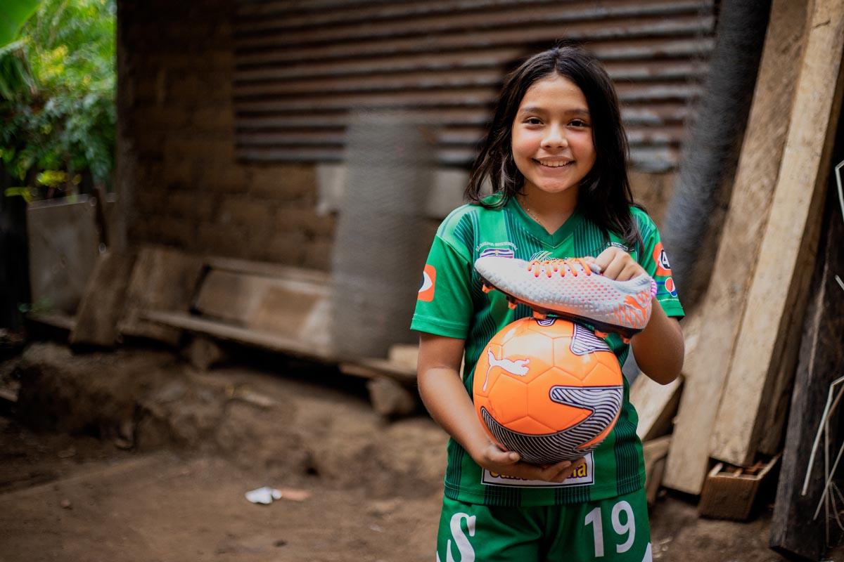 Karla, enfant parrainée par Compassion, se tient à l'extérieur de sa maison et tient un ballon de football orange et une paire de crampons.